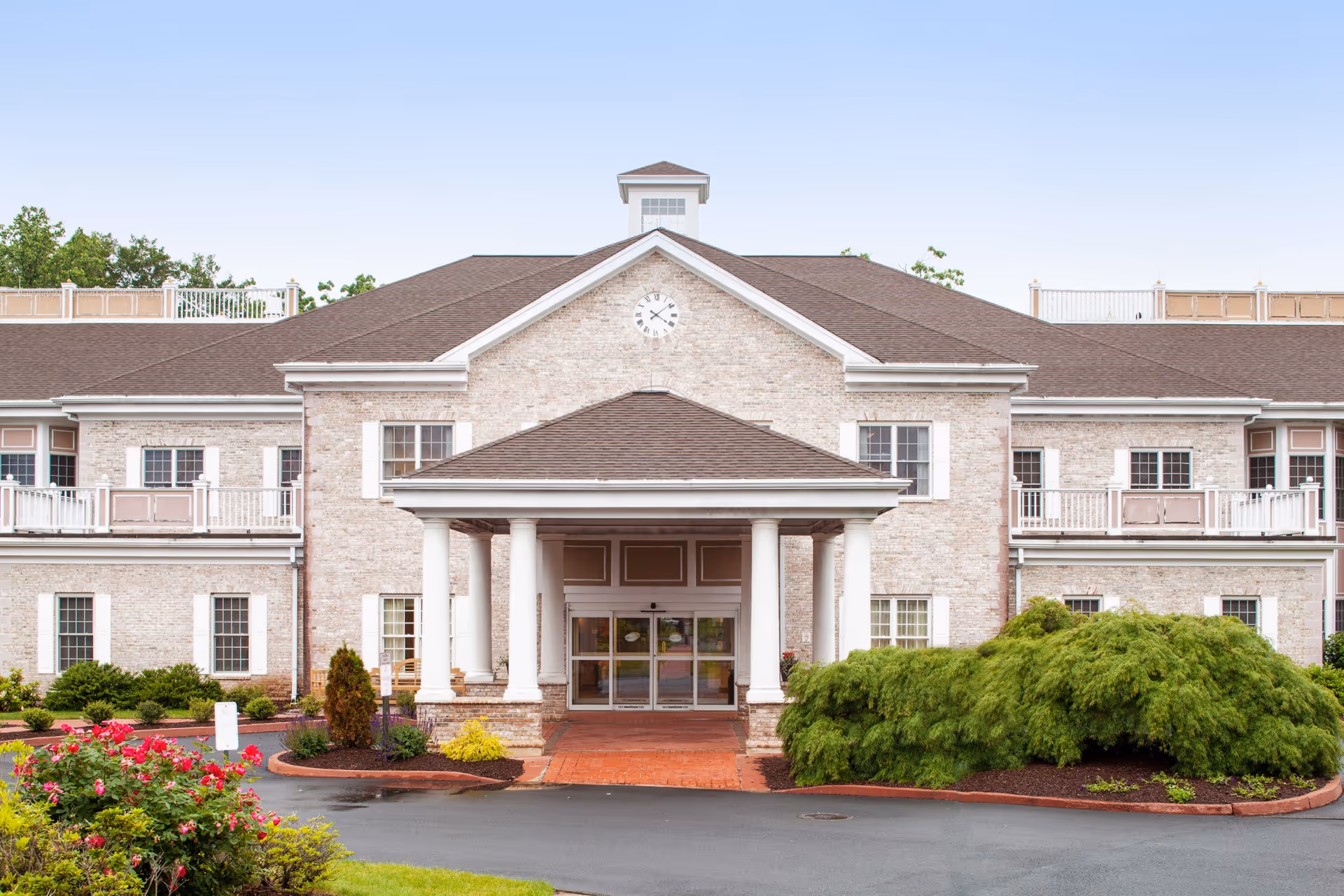 Front entrance of a two-story brick senior living facility with columns, balconies, and landscaped shrubs.