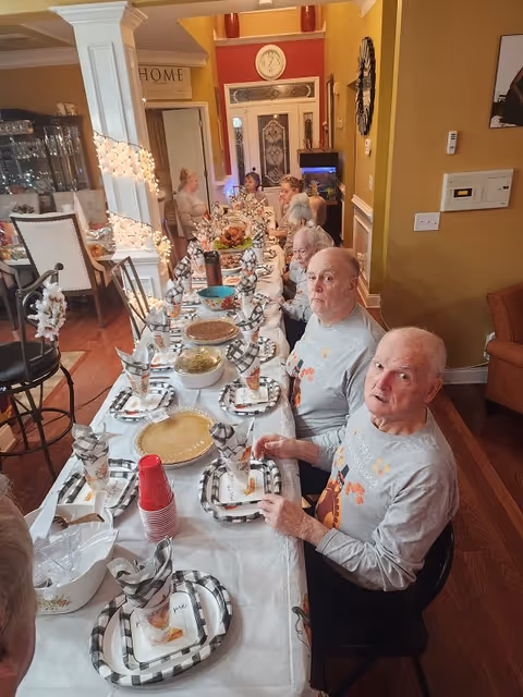 A group of elderly people seated around a long dining table set with plates, napkins, cups, and various dishes in a warmly lit dining room. The table is decorated with checkered napkins and there are festive lights wrapped around a pillar. The room has warm yellow walls and wooden flooring.