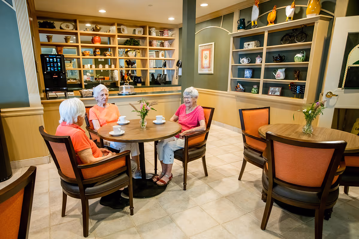 Three older adults sitting around a round table with cups in a bright communal dining area with shelves and decorative items behind them.