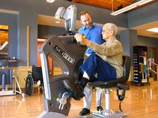An elderly man using a seated exercise bike in a fitness or rehabilitation area, assisted by a younger man standing beside him. The room has wooden floors and exercise equipment in the background.
