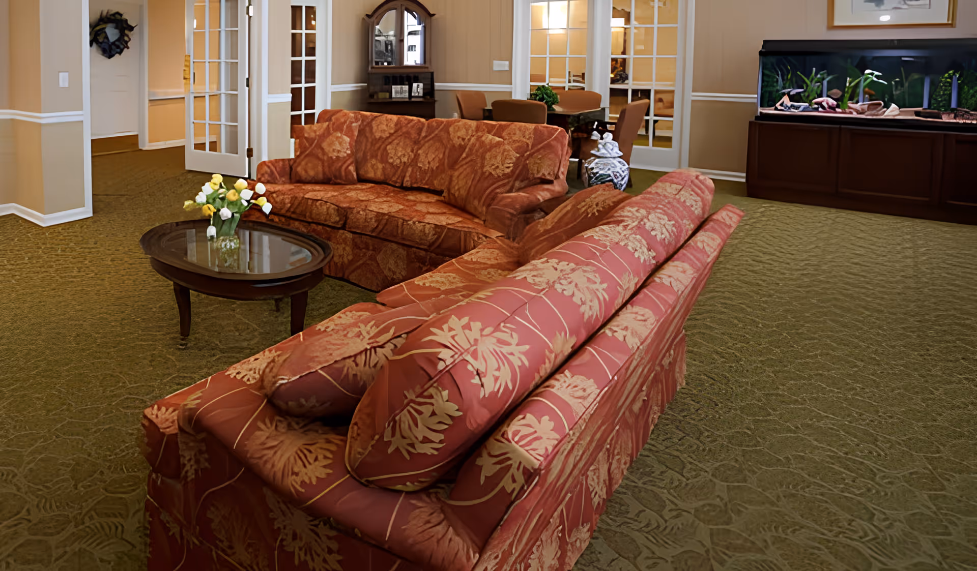 Furnished senior living common room with patterned sofas, a glass-top coffee table with flowers, dining chairs and a fish tank in the background.