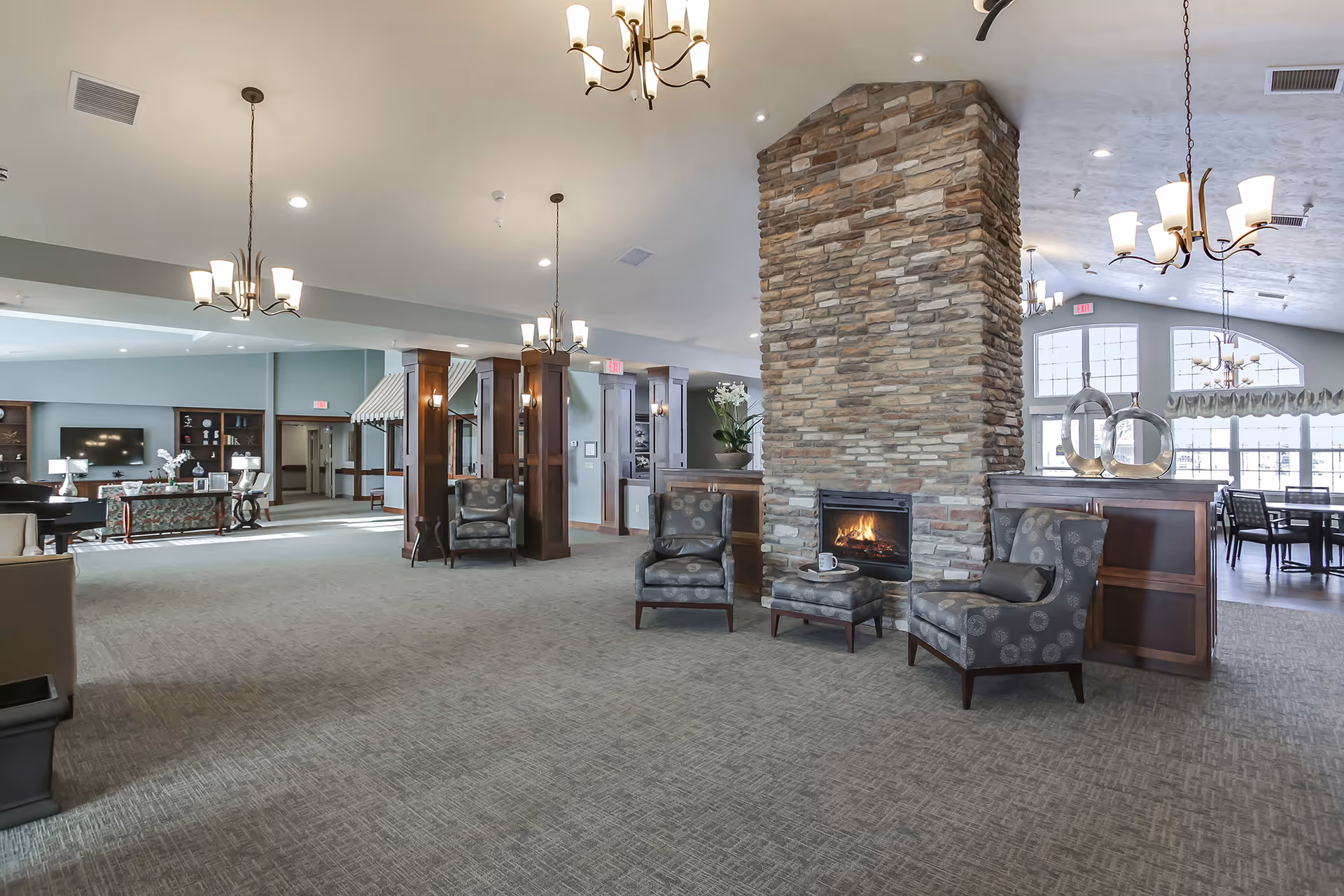 Spacious and well-lit common area in an assisted living facility featuring a large stone fireplace with a fire burning, surrounded by several patterned armchairs and an ottoman. The room has carpeted floors, multiple chandeliers hanging from the ceiling, large windows allowing natural light, and a dining area visible in the background.