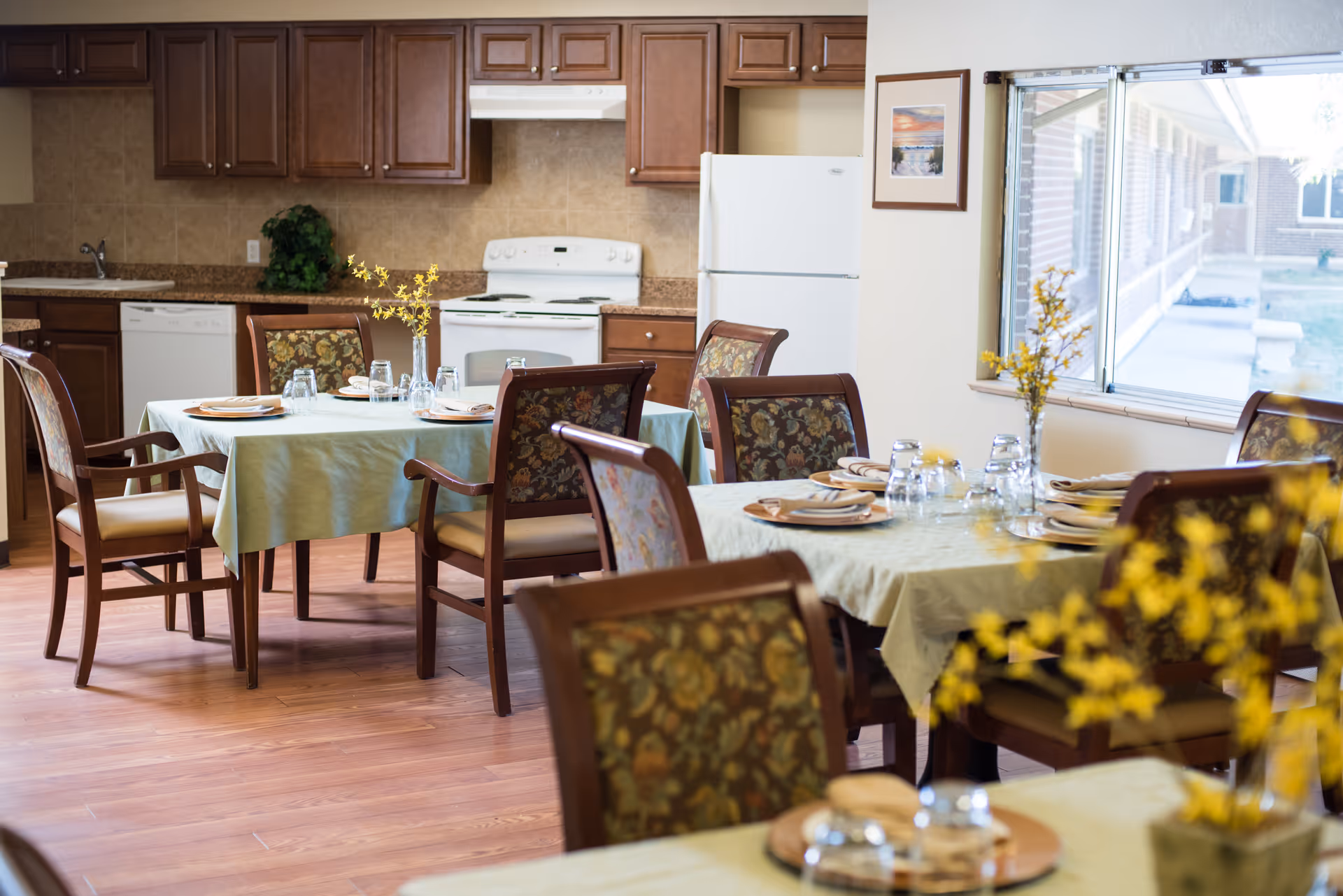 A dining area with tables covered in light green tablecloths, set with plates, glasses, and yellow floral centerpieces. The room has wooden chairs with floral upholstery and a kitchen area in the background featuring wooden cabinets, a white stove, refrigerator, and dishwasher. A large window on the right side lets in natural light and shows an outdoor walkway.