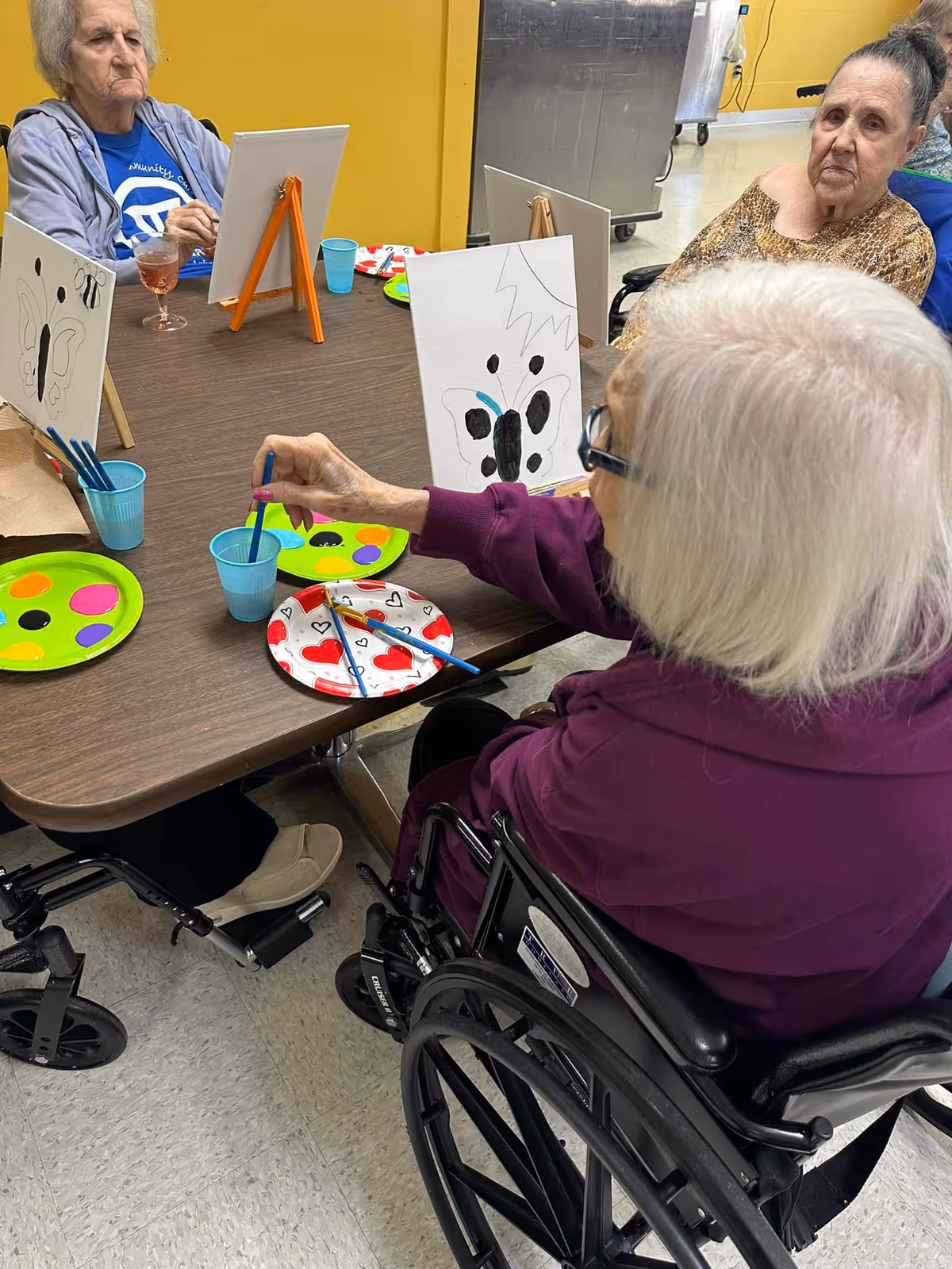 Three elderly women sitting around a table engaged in a painting activity. One woman in a wheelchair is painting a butterfly on a canvas, while the other two women watch. The table has paint palettes with bright colors, brushes, and cups of water. The room has a yellow wall and a tiled floor.