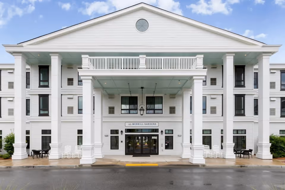 Front exterior view of a large white building with tall columns and a covered entrance. The building has multiple windows and a sign above the entrance that reads 'Merrill Gardens'. There are chairs and tables on either side of the entrance on the sidewalk.