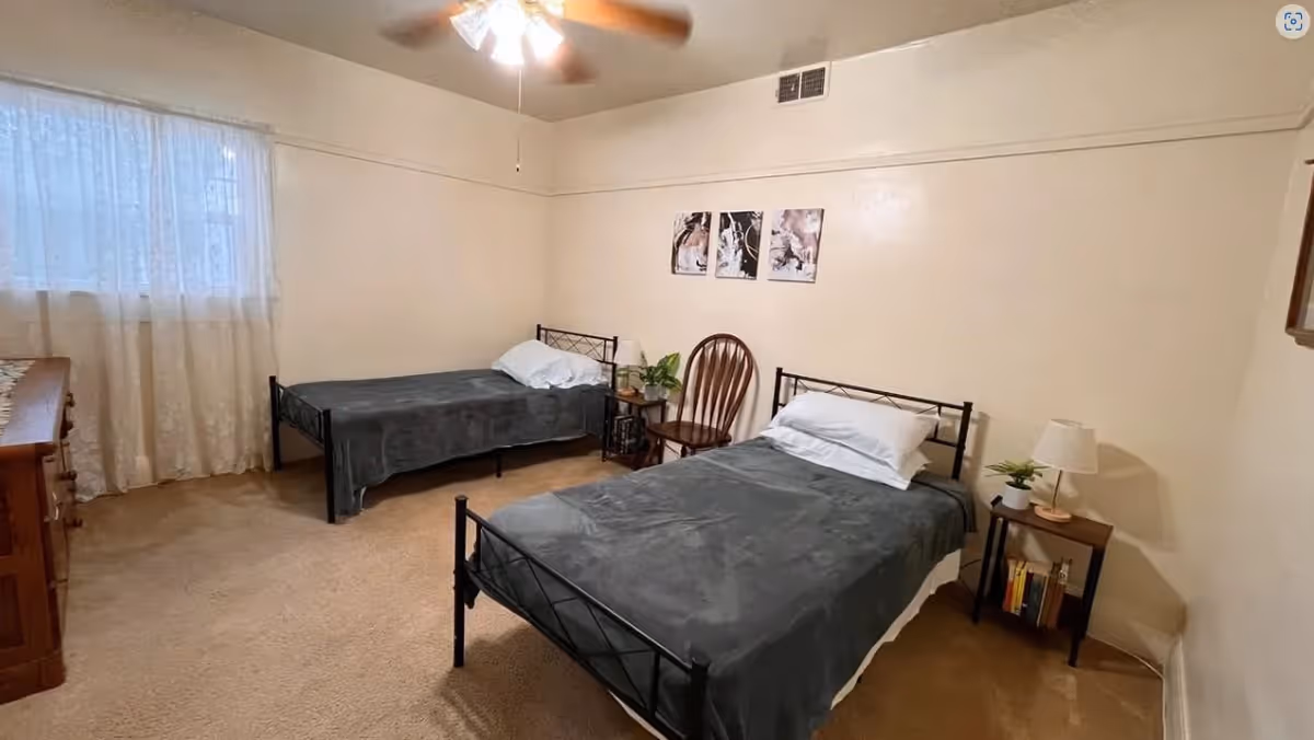 A simple bedroom with two single beds covered in dark gray blankets and white pillows. Between the beds is a wooden chair and a small side table with a plant and a lamp. Another small table with a lamp and books is next to the bed on the right. A wooden dresser is visible on the left side of the room. The walls are light-colored, and there is a window with sheer curtains on the left wall. Three abstract art pieces hang above the chair on the wall.