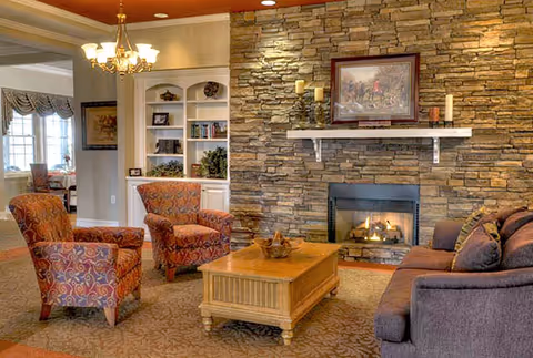 A cozy living room with a stone fireplace, a wooden coffee table, a purple sofa, and two patterned armchairs. There is a chandelier hanging from the ceiling and built-in white shelves with books and decorative items. A framed picture hangs above the fireplace.