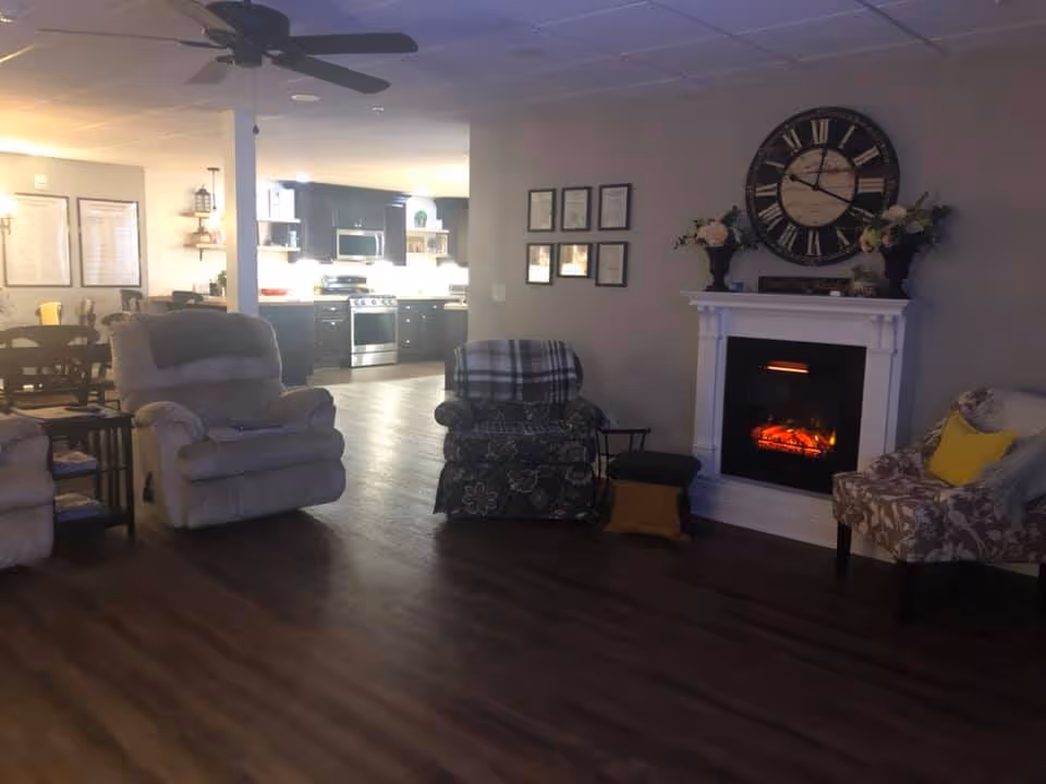 Cozy communal living room with recliners and armchairs arranged around an electric fireplace, open to a kitchen and dining area in the background.