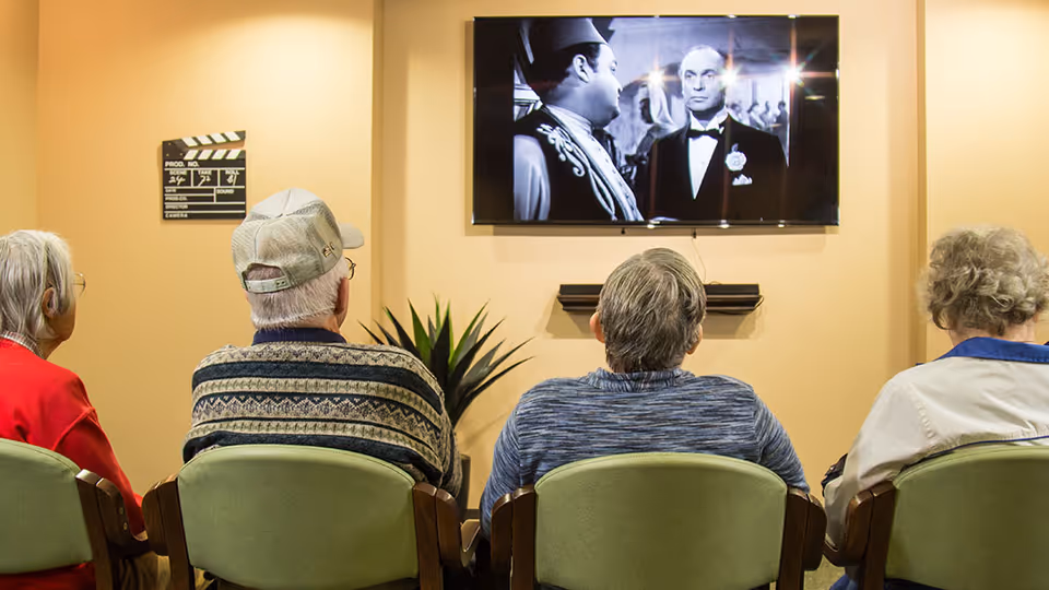 Four elderly people sitting in green chairs facing a wall-mounted television screen showing a black-and-white movie scene. The room has beige walls, a small plant, and a film clapperboard decoration on the wall.