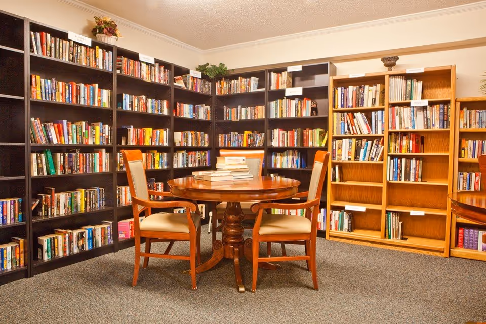 A cozy library room with bookshelves lining the walls and a round wooden table with chairs in the center.