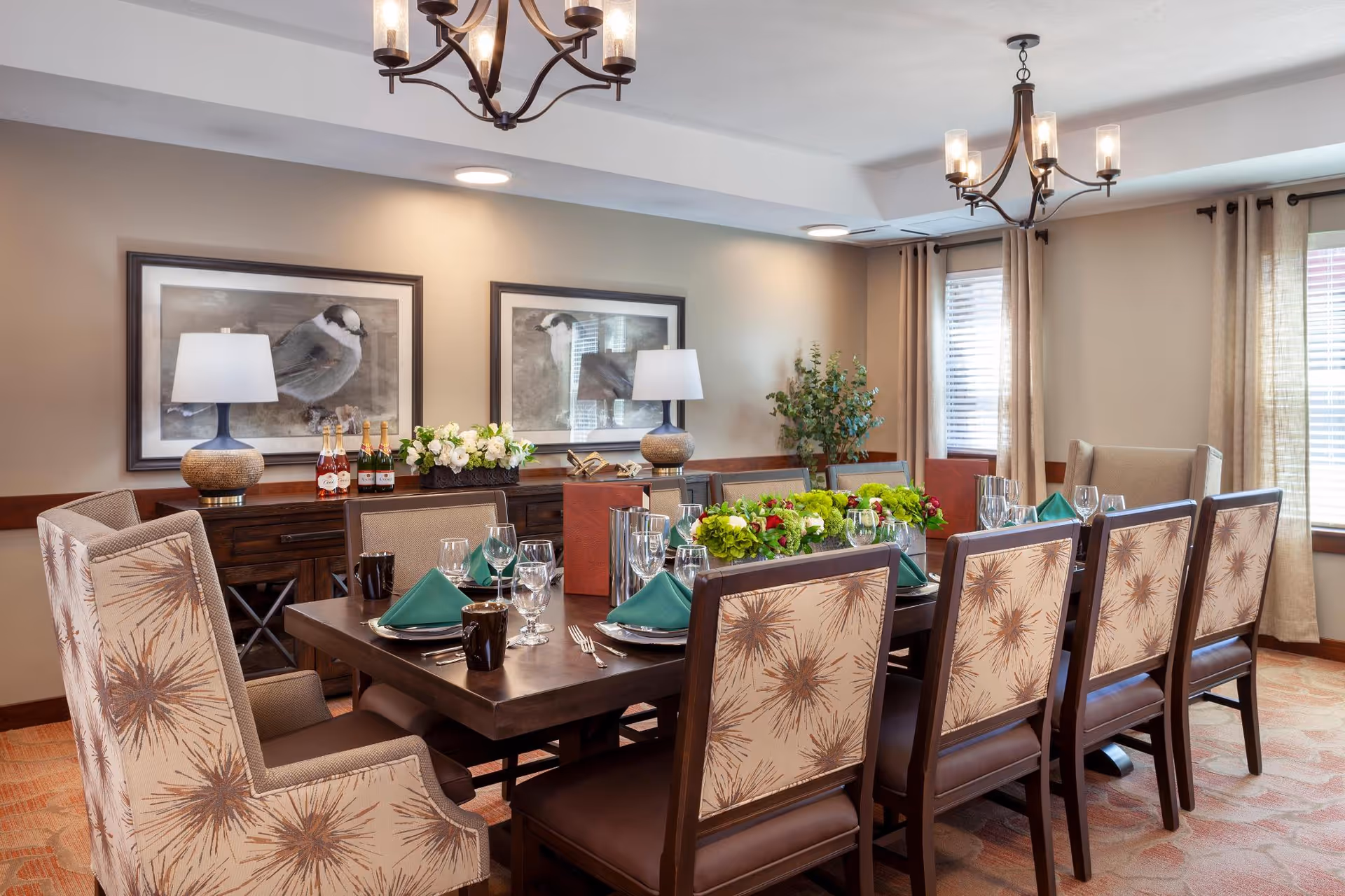 A well-decorated dining room with a long wooden table set for a meal. The table has green napkins, glassware, and floral centerpieces. Surrounding the table are upholstered chairs with a starburst pattern on the backrests. The room features two framed bird artworks on the wall, two table lamps on a sideboard, and large windows with beige curtains allowing natural light to enter.