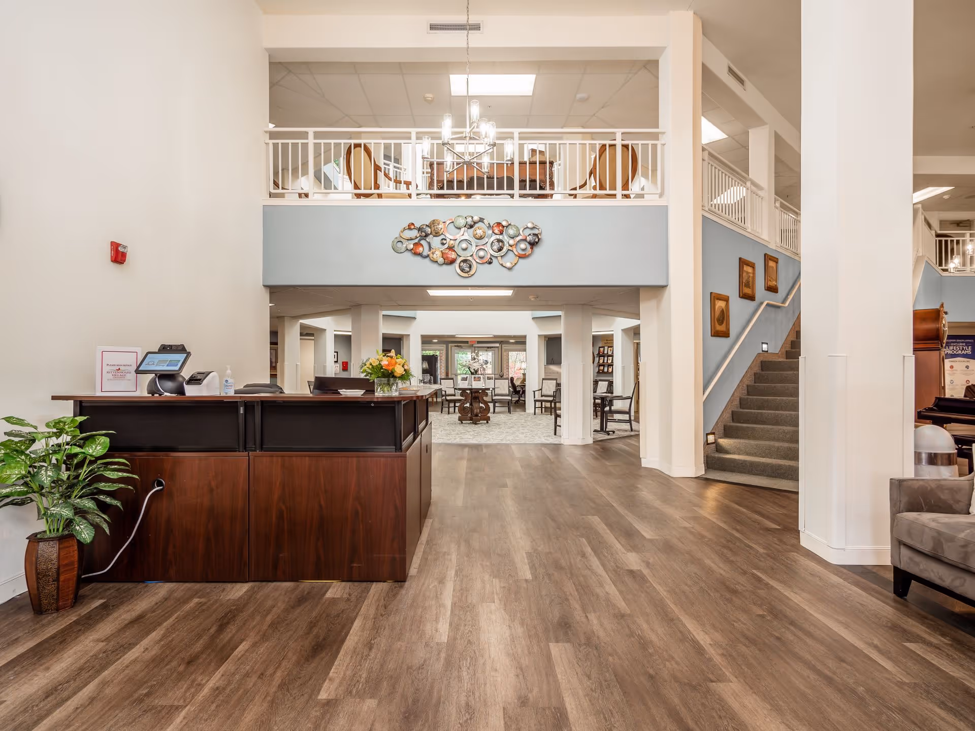 Spacious senior living lobby with a wooden reception desk, seating areas, staircase, upper balcony, and decorative wall art.