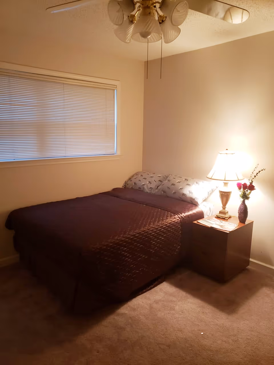 A simple bedroom with a made bed, a nightstand holding a lamp and vase of flowers, a window with closed blinds, and a ceiling fan.