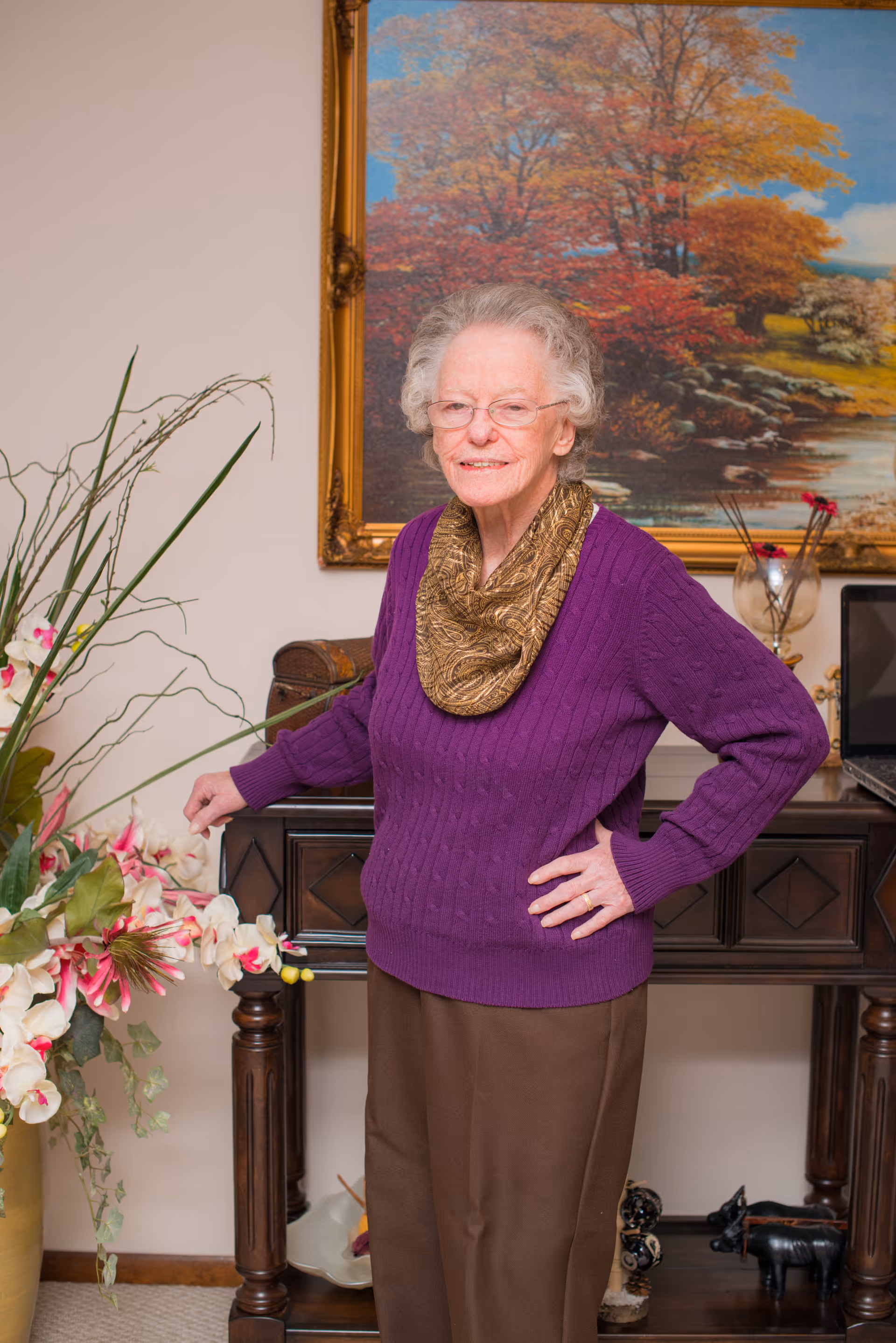 An elderly woman with white hair and glasses wearing a purple sweater and a gold scarf stands indoors next to a wooden table with decorative items and a large floral arrangement. Behind her is a framed painting of an autumn landscape with trees and a river.