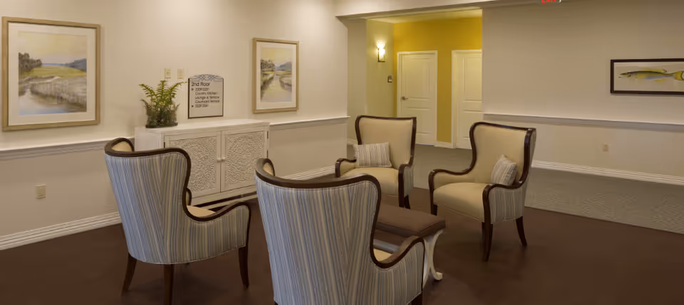 A bright lounge area with four upholstered armchairs arranged around a small table in a hallway-like common room.