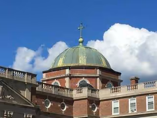 Exterior brick building with a green domed roof and cross against blue sky.