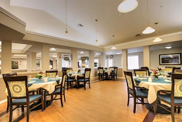 A spacious dining room in a senior living facility with multiple round tables covered with teal and beige tablecloths, each set with white napkins, glassware, and floral centerpieces. The room features wooden flooring, beige walls with white trim, several columns, pendant lighting, and large windows allowing natural light.