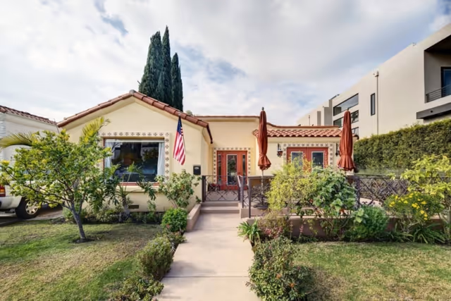 Front view of a single-story stucco house with a red tile roof, an American flag, a central walkway, and a small fenced patio with umbrellas.
