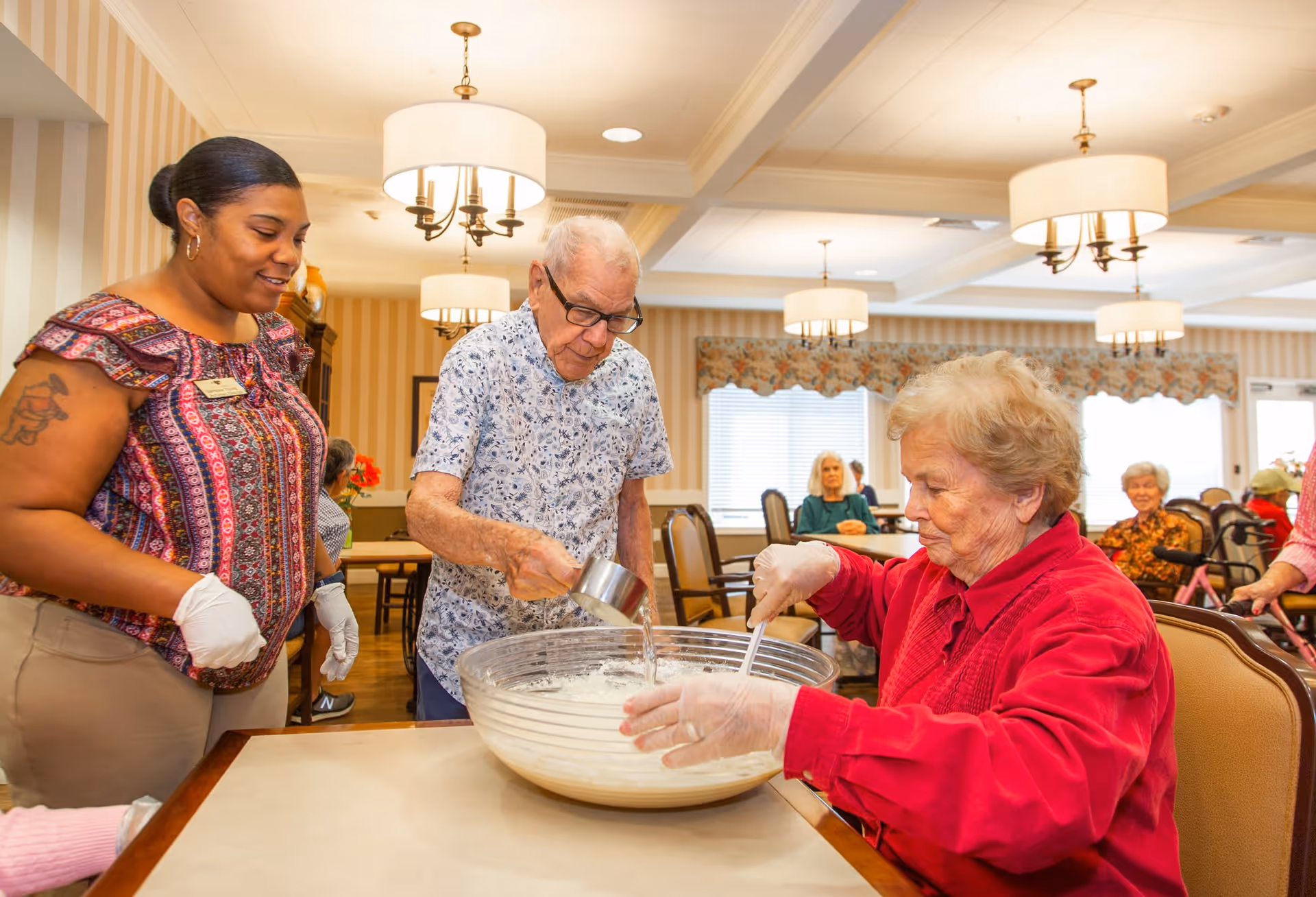 An elderly man and woman wearing gloves are mixing ingredients in a large glass bowl on a table, while a staff member watches and smiles in a well-lit dining room with other elderly residents seated in the background.