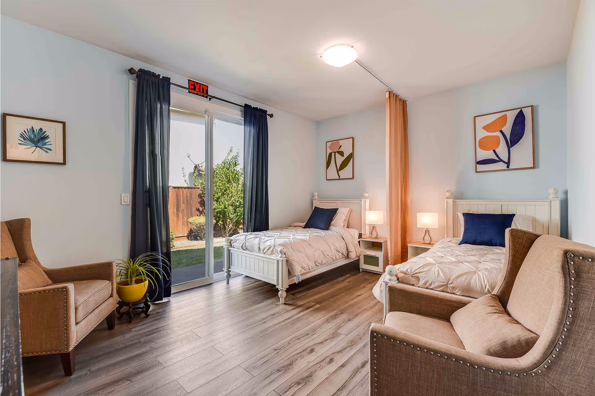 A bright and cozy bedroom with two single beds separated by a peach-colored privacy curtain. Each bed has white bedding and a dark blue pillow. There are two bedside tables with lamps between the beds. The room features light wood flooring, two beige armchairs, framed botanical artwork on the walls, and a sliding glass door with dark curtains leading to an outdoor garden area.