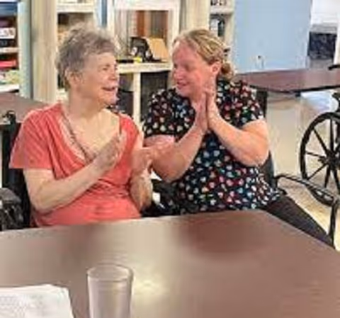 Two women sitting at a table in a communal indoor setting, smiling and clapping their hands. One woman is wearing a coral-colored top and the other is wearing a black top with a colorful heart pattern. There are wheelchairs and tables visible in the background.