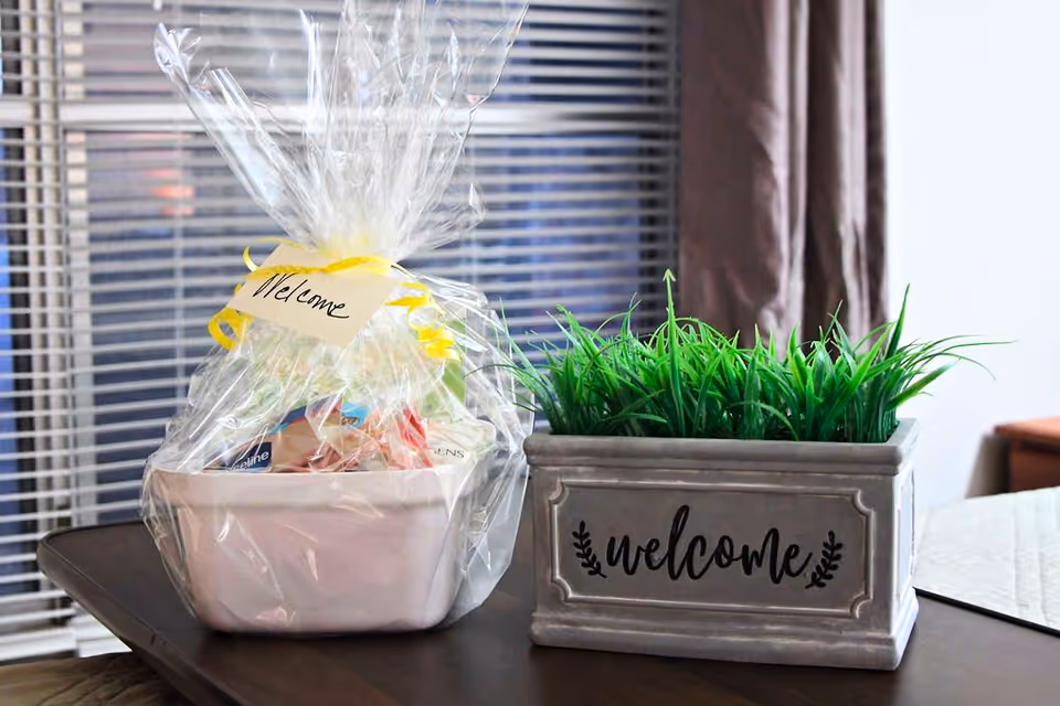 A welcome gift basket wrapped in clear plastic with a yellow ribbon and a handwritten 'Welcome' tag, placed on a table next to a rectangular planter with green artificial grass and the word 'welcome' written on it. In the background, there are window blinds and a curtain.