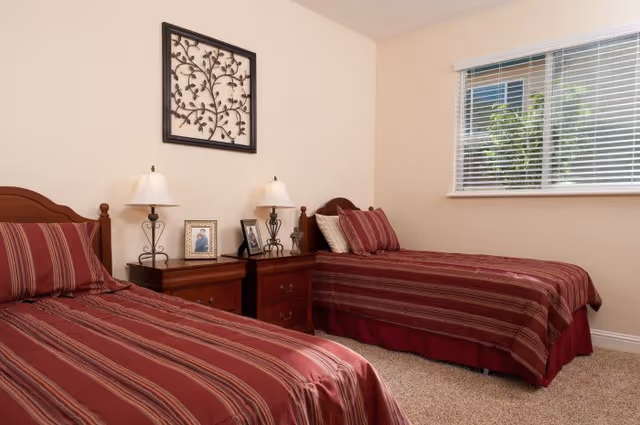 Tidy bedroom with two twin beds in red striped bedding, a shared nightstand with lamps and framed photos, and a window with blinds.