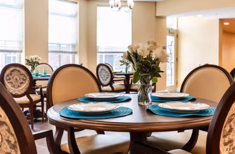 A dining room with a round wooden table set with white plates on teal placemats and a vase of white roses in the center. The room has multiple windows with blinds and several chairs with patterned upholstery around other tables in the background.