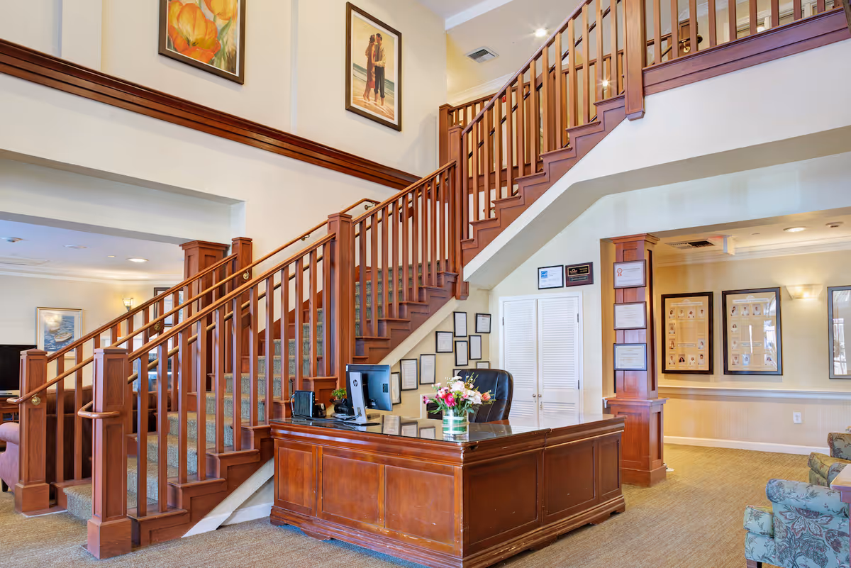 Interior view of a senior living facility lobby area featuring a wooden reception desk with a computer and a vase of flowers. Behind the desk is a staircase with wooden railings leading to an upper floor. The walls are decorated with framed certificates and artwork, and there is comfortable seating visible to the right.