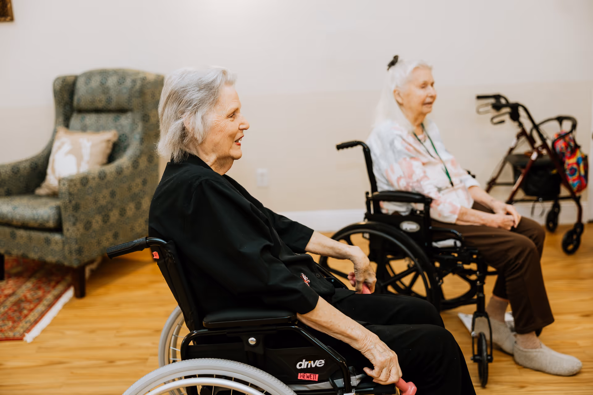 Two elderly women sitting in wheelchairs in a room with wooden flooring. One woman is wearing a black outfit and holding pink hand weights, while the other woman is dressed in a floral top and brown pants. Behind them is a green patterned armchair with a decorative pillow and a walker with a colorful bag hanging on it.