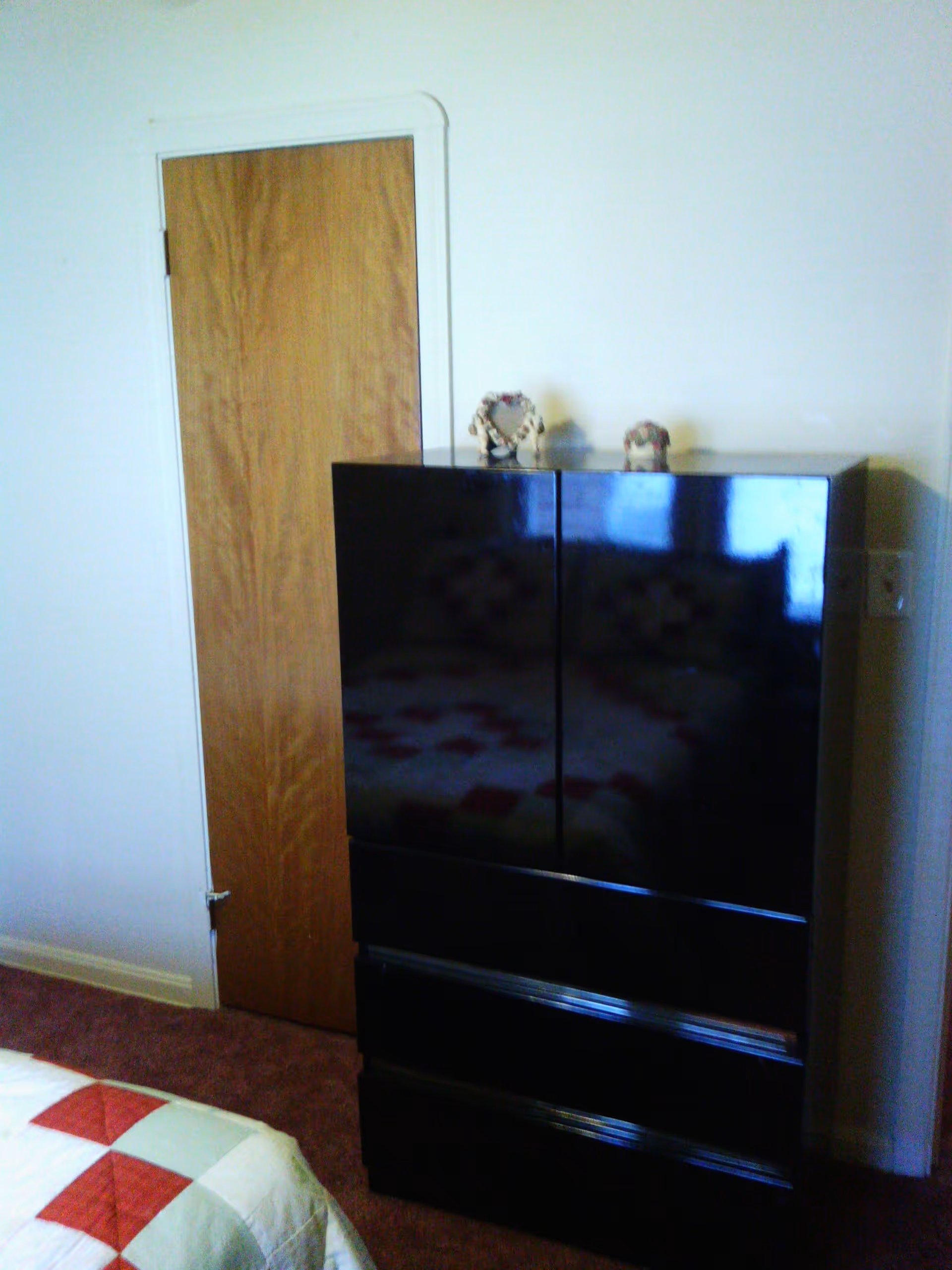 Corner of a bedroom featuring a wooden door, a glossy black dresser, and the corner of a quilted bed.