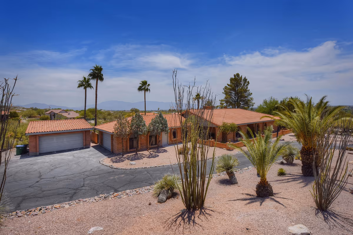 Exterior view of a single-story building with a red tile roof surrounded by desert landscaping including palm trees and cacti under a blue sky with some clouds.