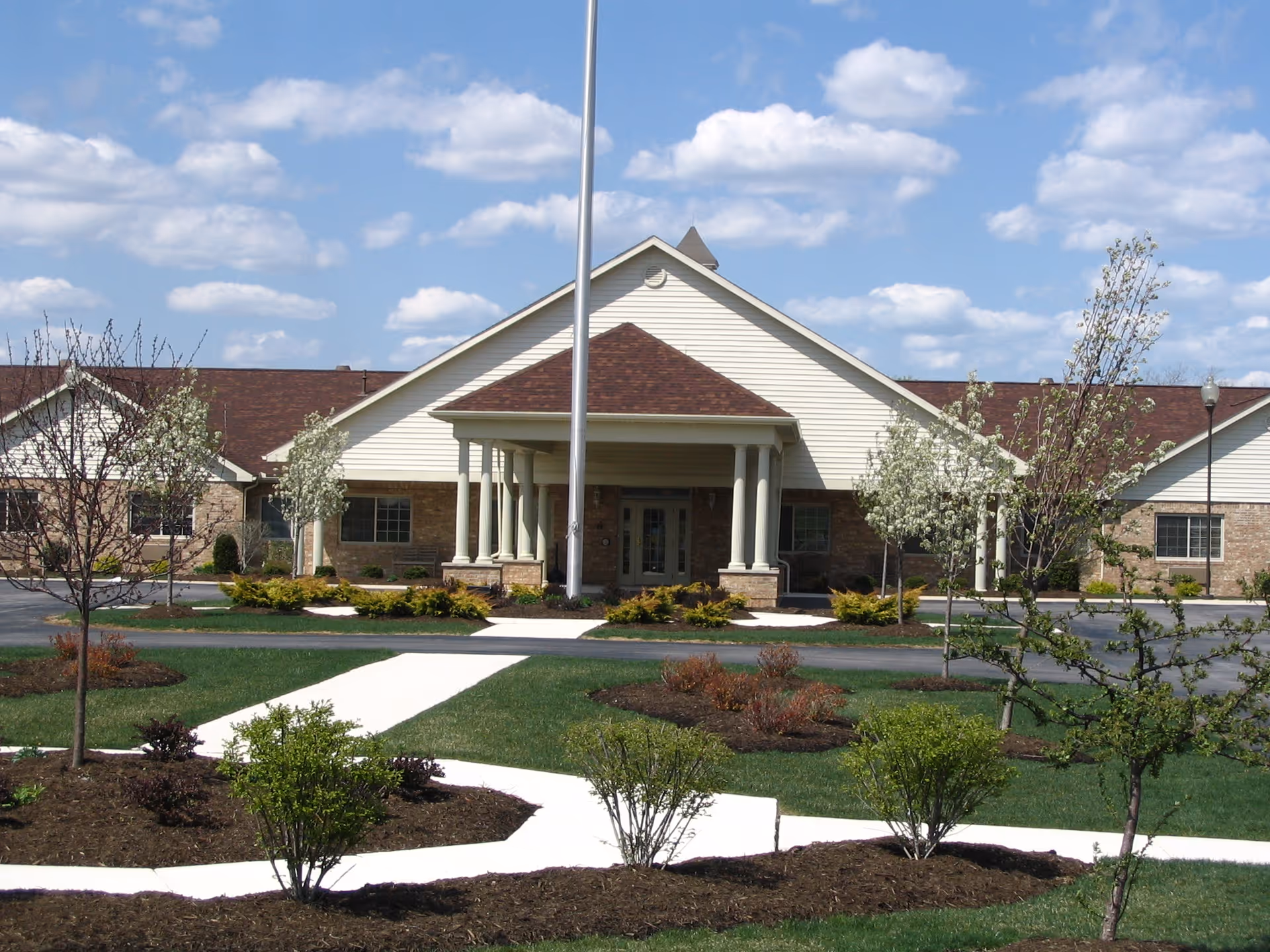 Front exterior view of a single-story senior living facility building with a peaked roof and a covered entrance supported by columns. The building is surrounded by landscaped gardens with small trees and shrubs, and a concrete walkway leads to the entrance. The sky is partly cloudy.
