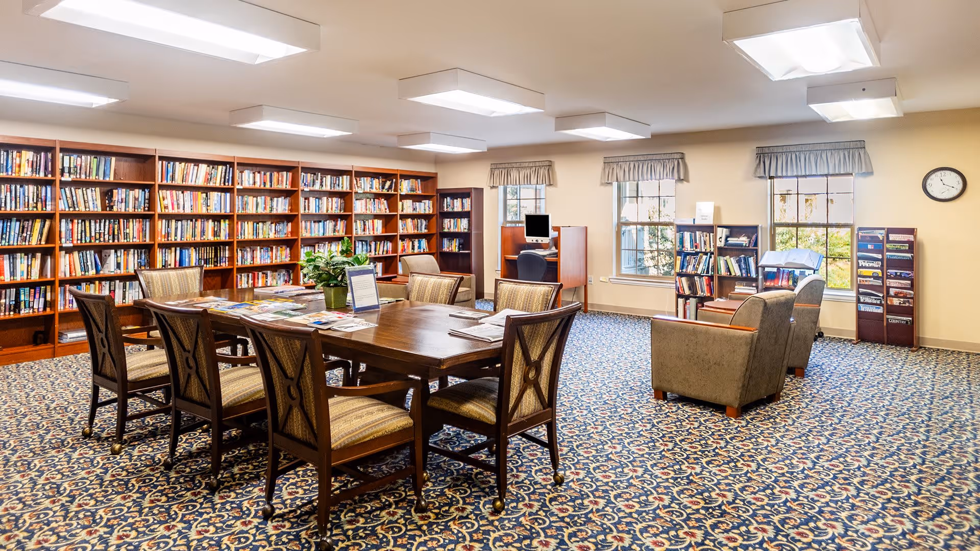A spacious library room with wooden bookshelves filled with books along the walls, a large wooden table with six chairs in the center, two armchairs near the windows, a computer workstation, and a patterned carpet covering the floor.
