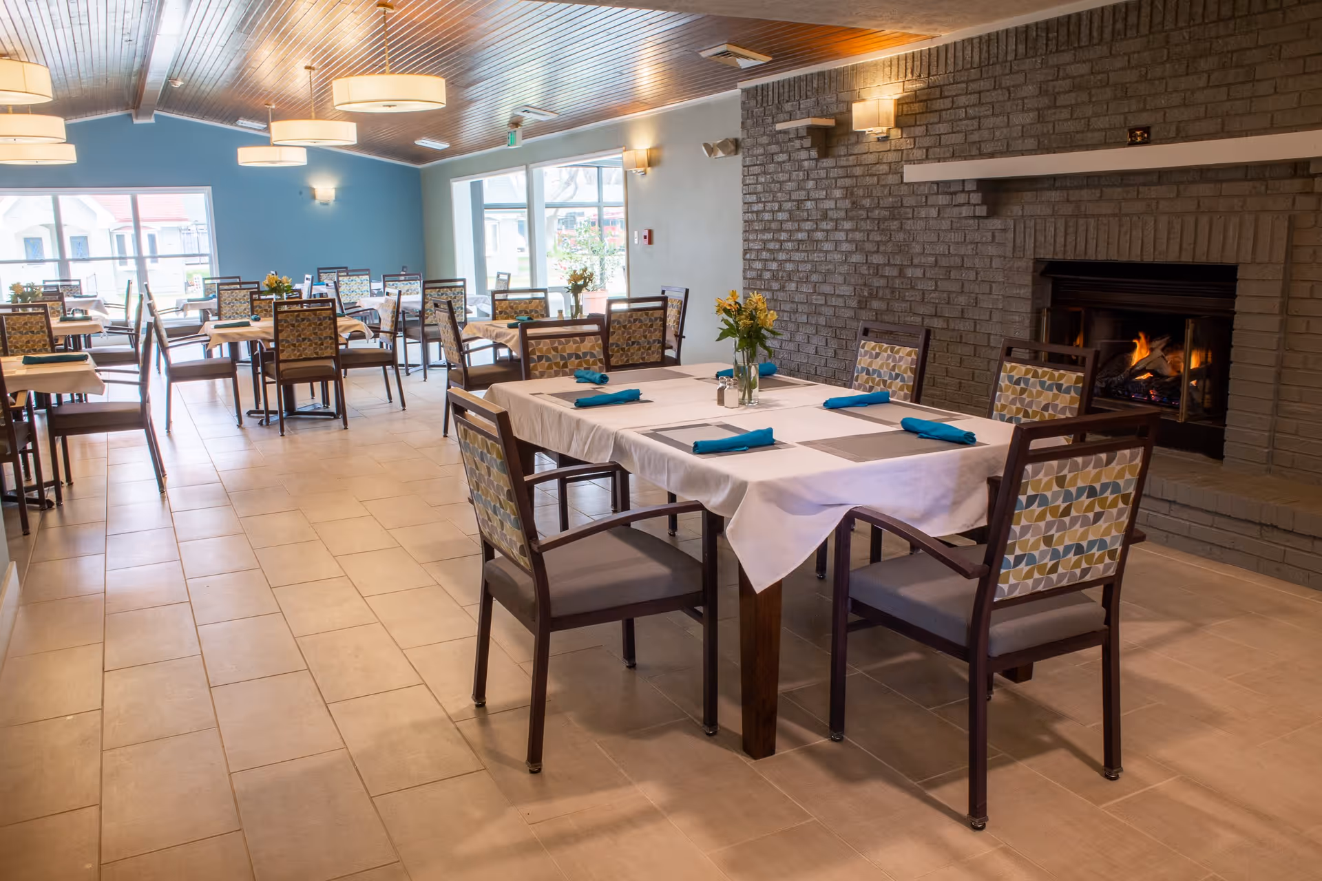 A spacious dining room with multiple tables covered in white tablecloths and set with blue napkins. Chairs with patterned backs surround the tables. A lit fireplace is built into a gray brick wall on the right side. Large windows on the left let in natural light, and modern ceiling lights hang from a wooden ceiling.