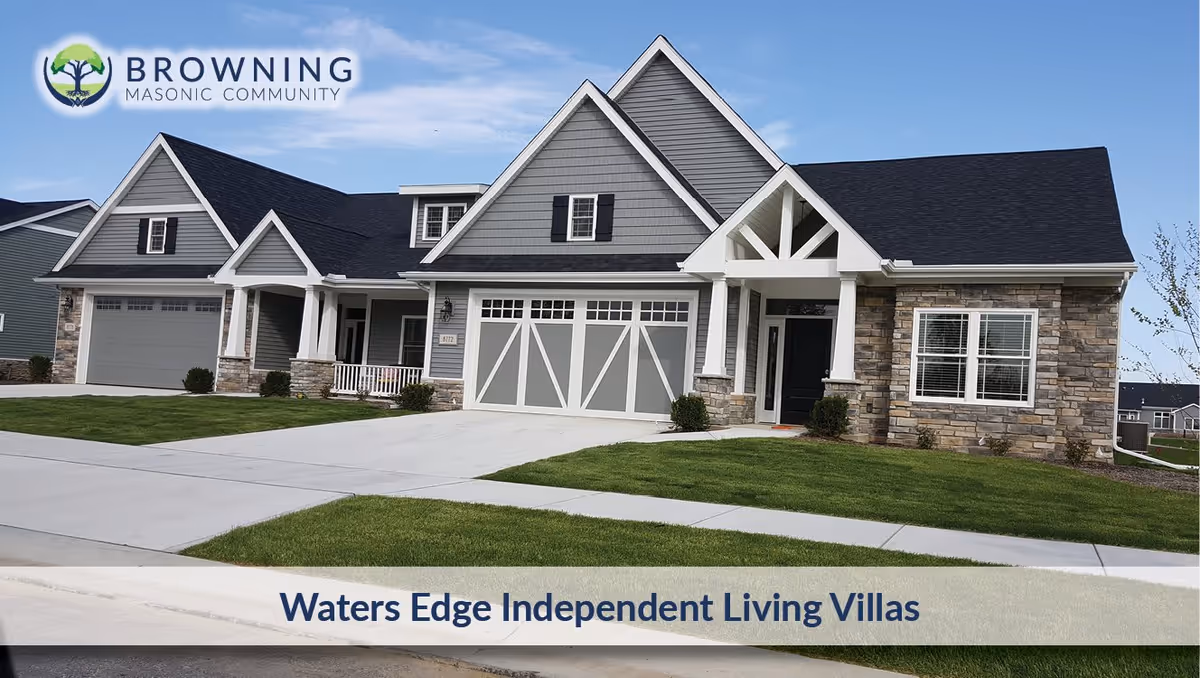 Exterior view of a modern single-story villa with gray siding, stone accents, a two-car garage, and a well-maintained lawn under a clear blue sky. The image includes the Browning Masonic Community logo and the text 'Waters Edge Independent Living Villas'.