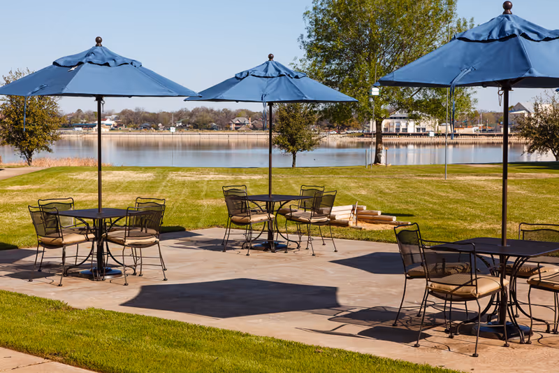 Outdoor patio area with three black metal tables, each with four chairs and blue umbrellas providing shade. The patio overlooks a grassy lawn and a body of water with trees and houses in the background under a clear blue sky.