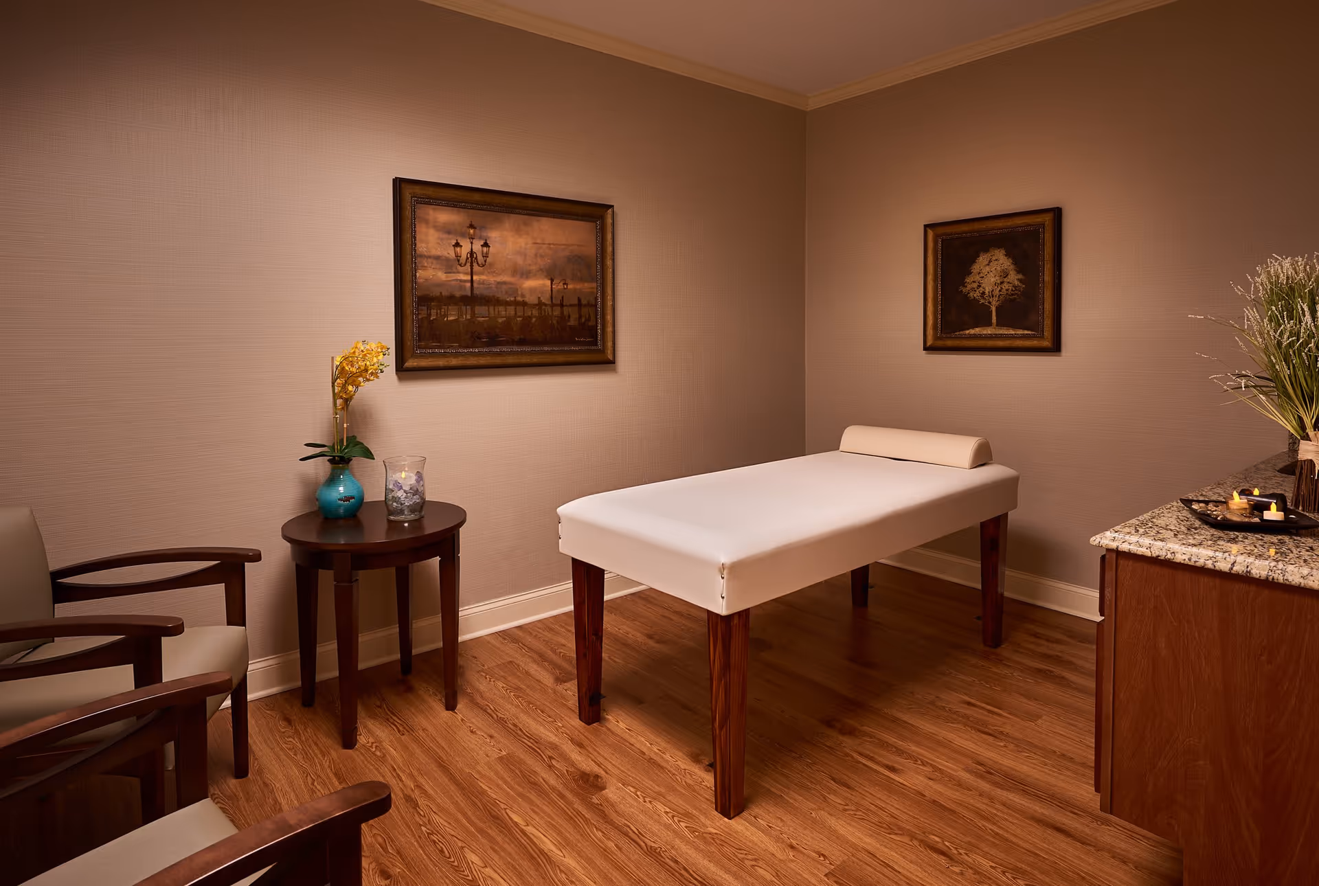 A warm-toned therapy room with a massage table, two chairs, a small side table with a vase, and framed artwork on the walls.