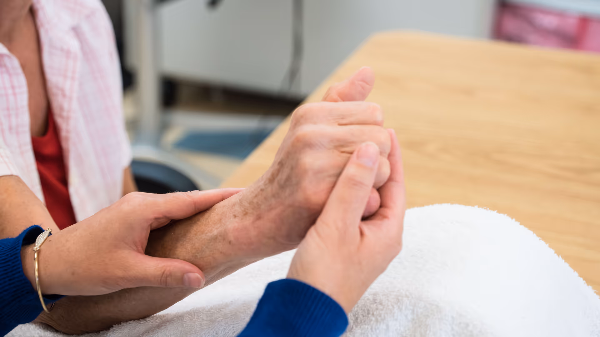 Close-up of a caregiver holding and gently supporting an elderly person's hand and wrist over a white towel on a wooden table.