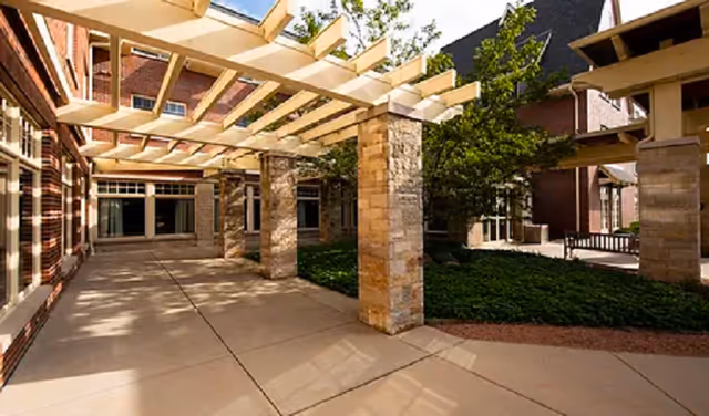 Sunlit outdoor courtyard with a wooden pergola, stone columns, paved walkways and adjacent brick building.