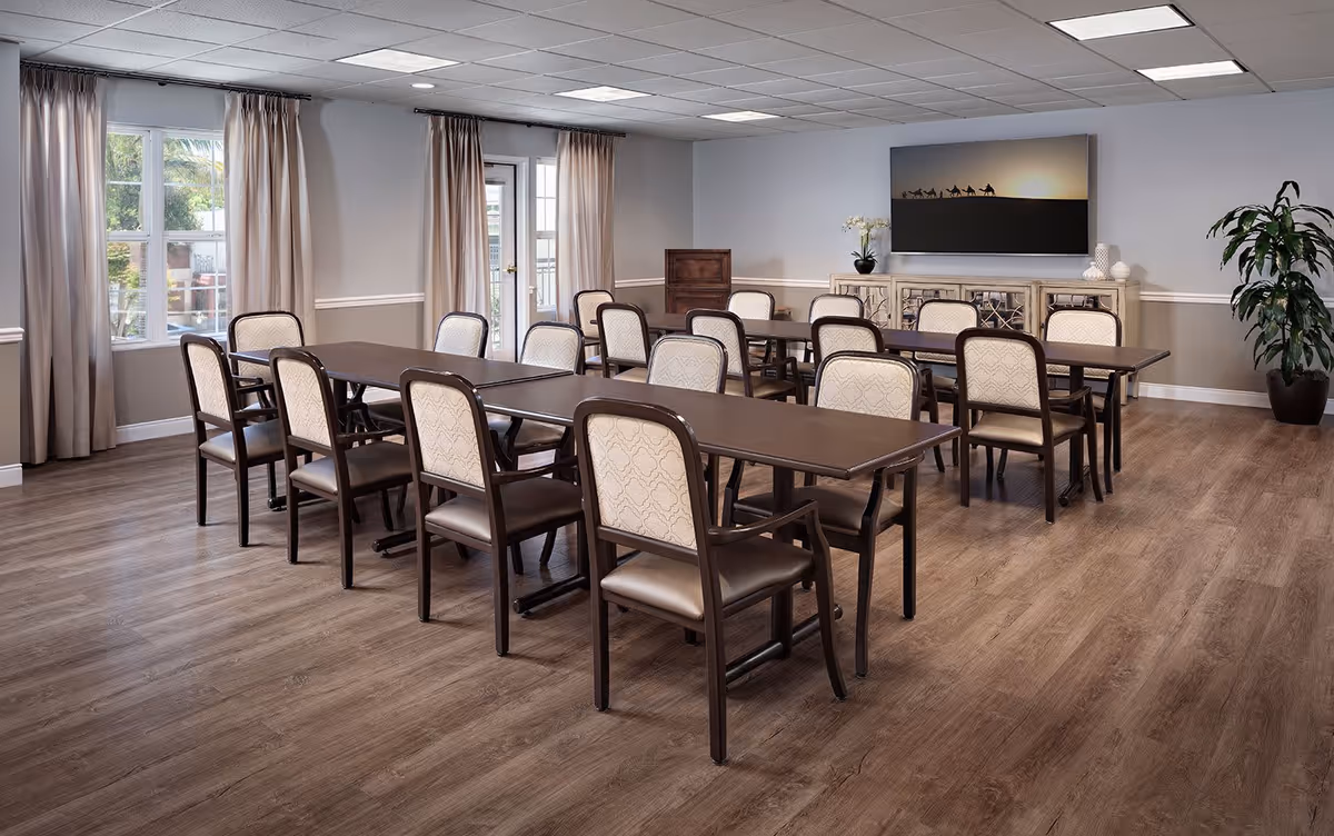 A well-lit dining room with two long rectangular tables and several cushioned chairs arranged around them. The room has wood flooring, beige walls with white trim, large windows with beige curtains, a door with glass panels, a decorative sideboard with a plant and vases, a large framed picture of camels on a desert landscape, and a tall potted plant in the corner.