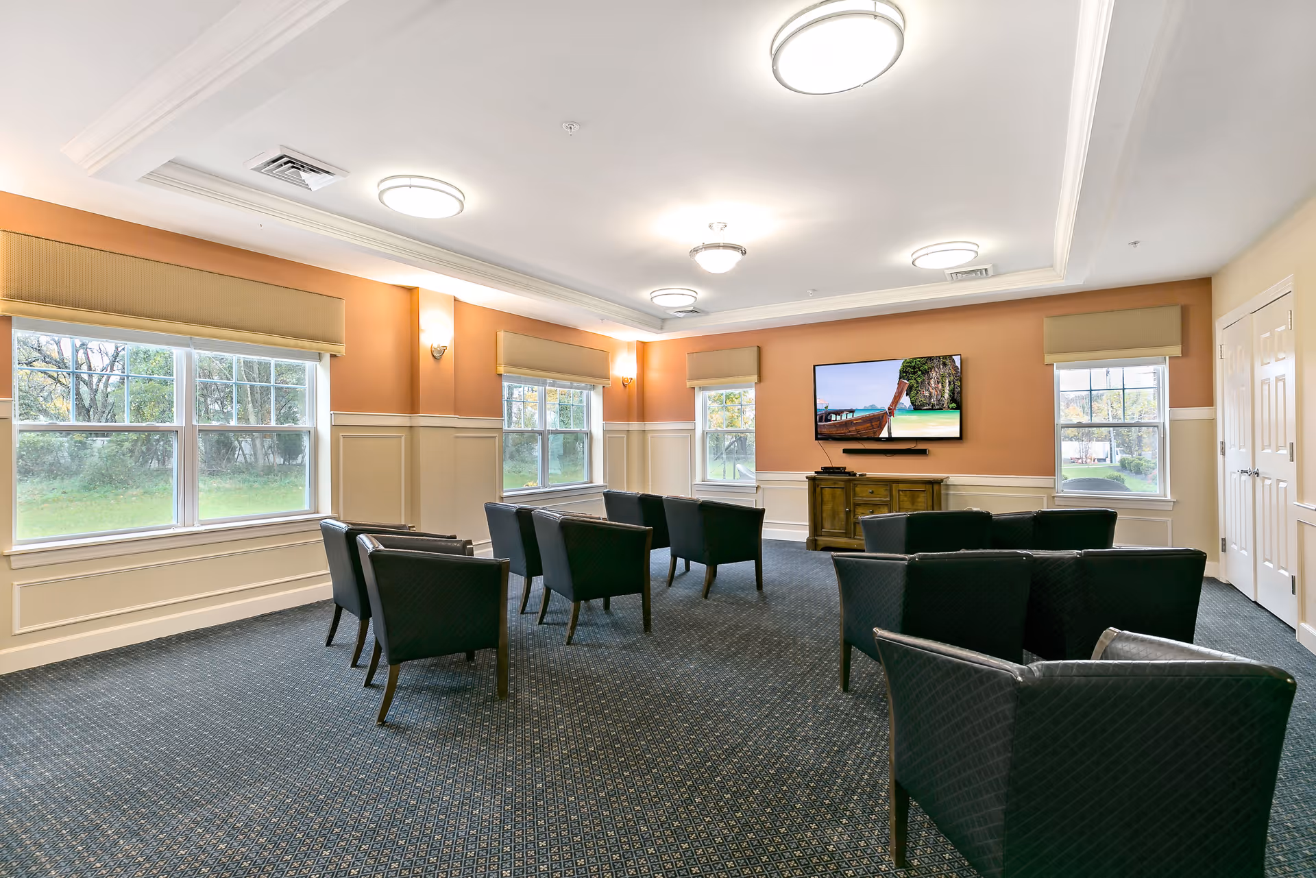 A well-lit common room with multiple black armchairs arranged in rows facing a wall-mounted flat-screen TV. The room has large windows with beige valances, peach and cream colored walls, and a patterned carpet. Ceiling lights provide additional illumination.