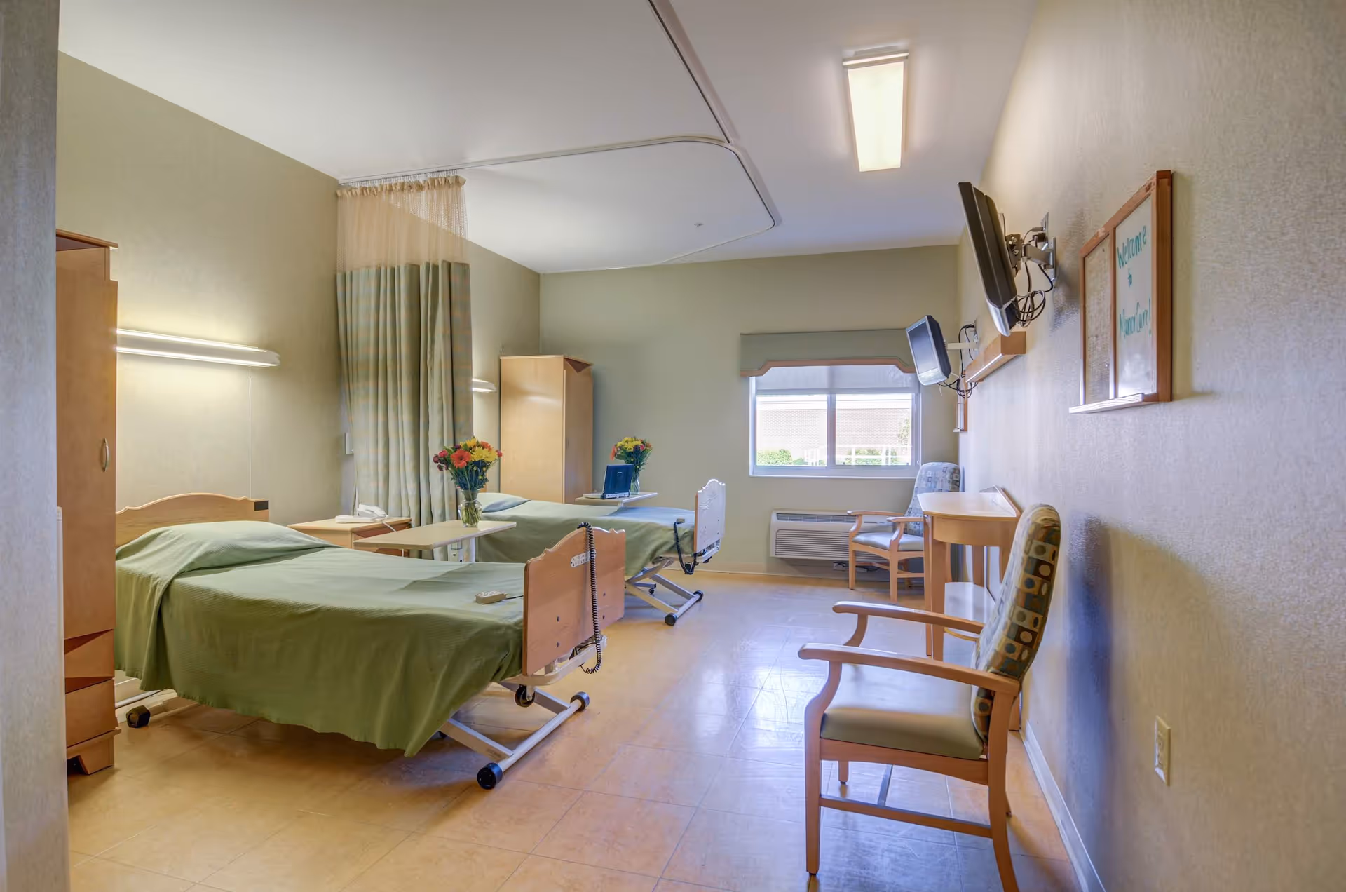 A shared senior care bedroom with two green-covered adjustable beds, bedside tables with flowers, chairs, a wall-mounted TV, and a window.