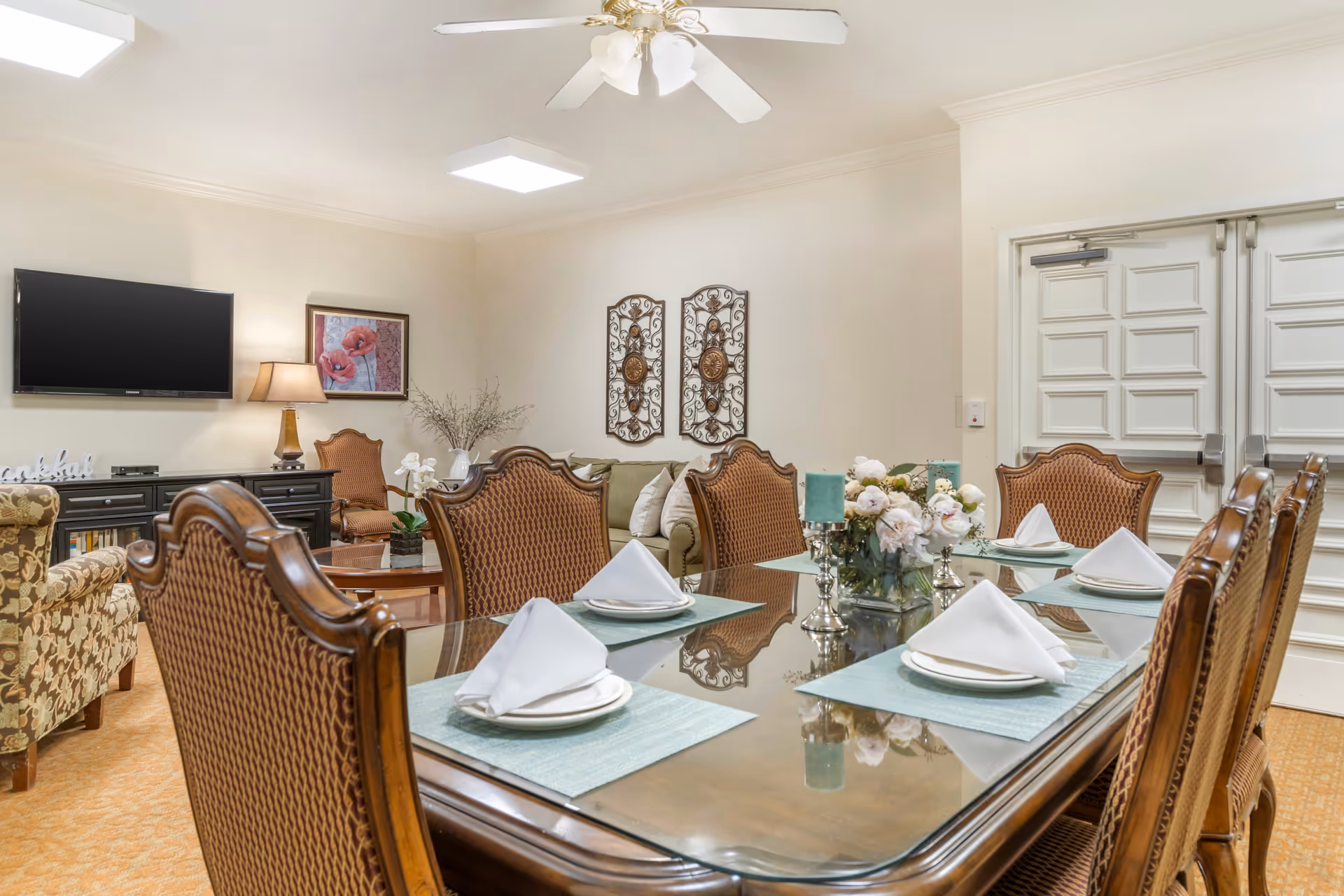 A well-decorated dining room with a large glass-top wooden table set for six with white napkins on plates and light blue placemats. The room features ornate wooden chairs, a floral centerpiece, and decorative candles. In the background, there is a living area with a green couch, a floral armchair, a wall-mounted TV, a table lamp, and wall art. The walls are light-colored, and there is a ceiling fan with lights above.