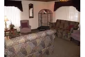 A living room with patterned sofas and two purple armchairs. There is a small table with a lamp and decorative items, a mirror on the wall, and a metal shelving unit in the corner. The room has carpeted flooring and windows with white curtains and dark valances.