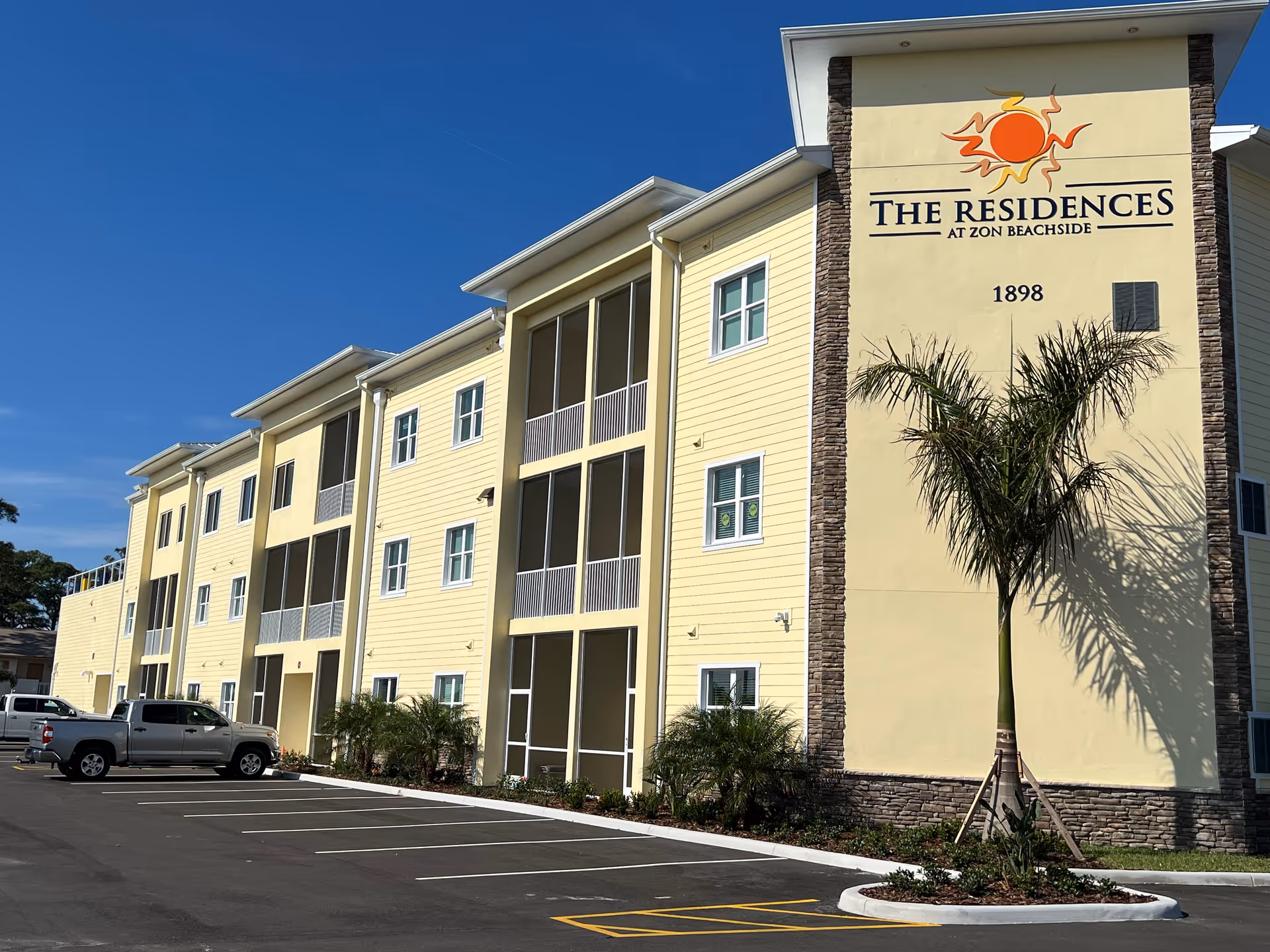 Exterior view of a three-story yellow building with multiple windows and screened balconies. The building has a sign that reads 'The Residences at Zon Beachside 1898' with a sun logo above it. There are palm trees and a parking lot with several parked vehicles in front of the building under a clear blue sky.