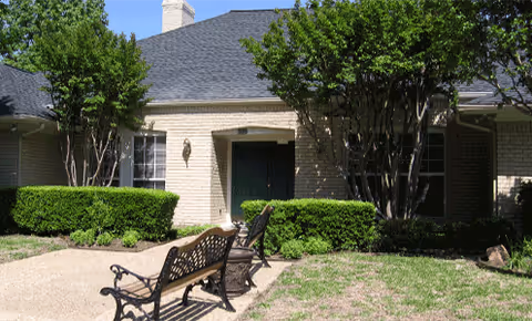 Exterior view of a single-story brick building with a dark shingled roof, surrounded by trimmed bushes and trees. There is a paved walkway leading to a dark green door, and two black metal benches are placed on the walkway in front of the entrance.