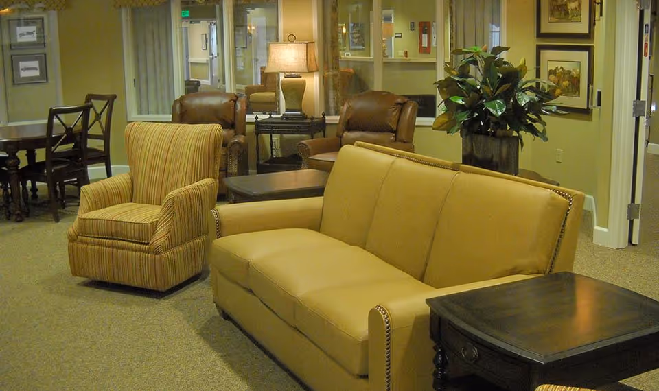 A cozy assisted living common room with a tan sofa, striped armchair, leather recliners, side tables, a lamp, and a potted plant.