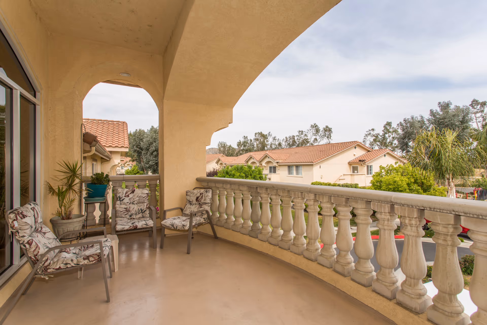 Curved outdoor balcony with beige stucco walls and a balustrade railing, furnished with four cushioned chairs and potted plants, overlooking a residential area with tiled roofs and trees under a cloudy sky.