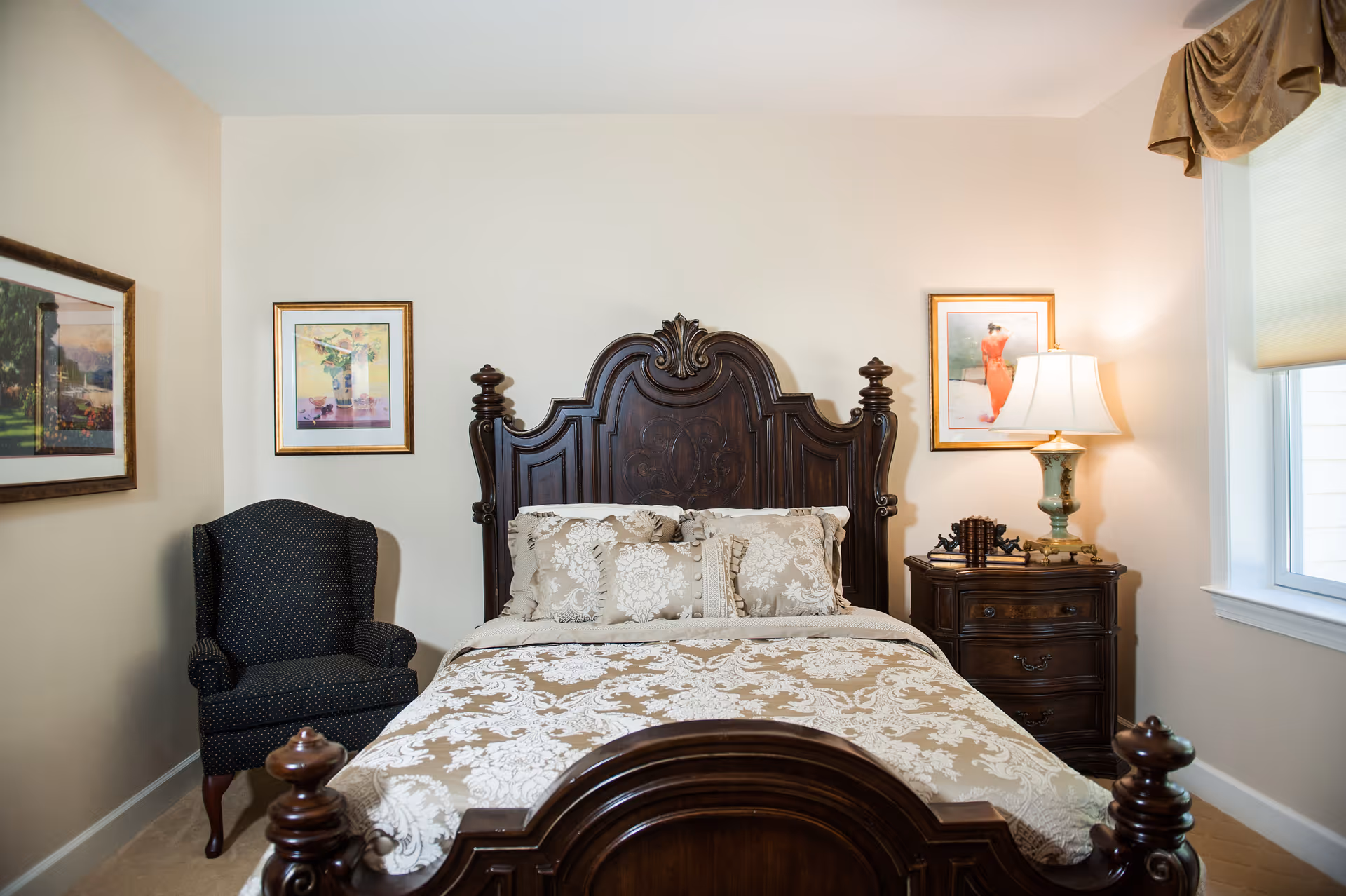 A bedroom with a dark wooden ornate bed frame and matching nightstand. The bed is made with a beige and white floral patterned comforter and pillows. There is a dark upholstered armchair to the left of the bed. Two framed pictures hang on the walls above the nightstand and armchair. A table lamp with a white shade sits on the nightstand next to a window with a beige valance.